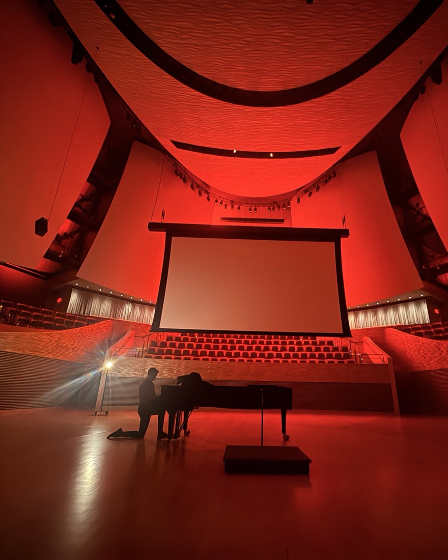 Nate playing a grand piano alone on the stage of a concert hall lit in red